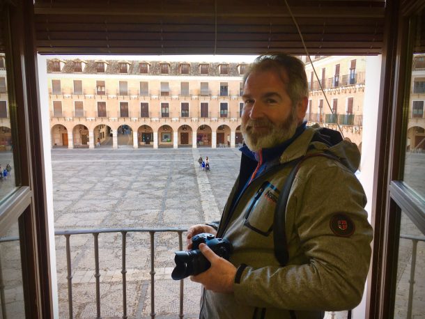 A man holding a camera and wearing a coat smiles in front of a town square