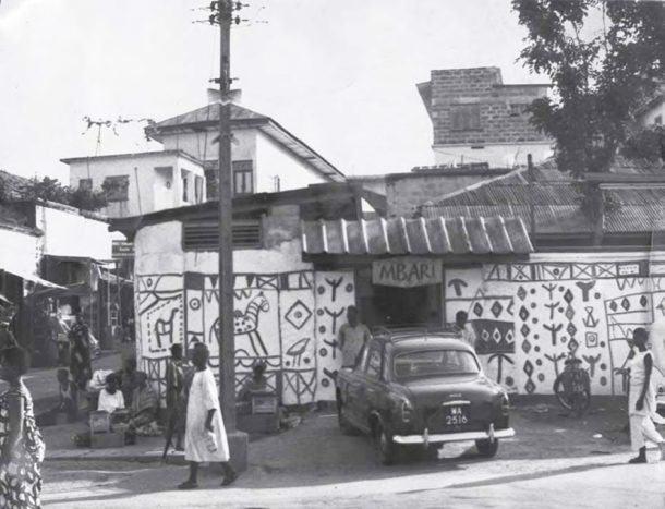 Black and white photograph of a white building with patterns painted onto it (including a zebra). A car is outside the building and people are walking along the street