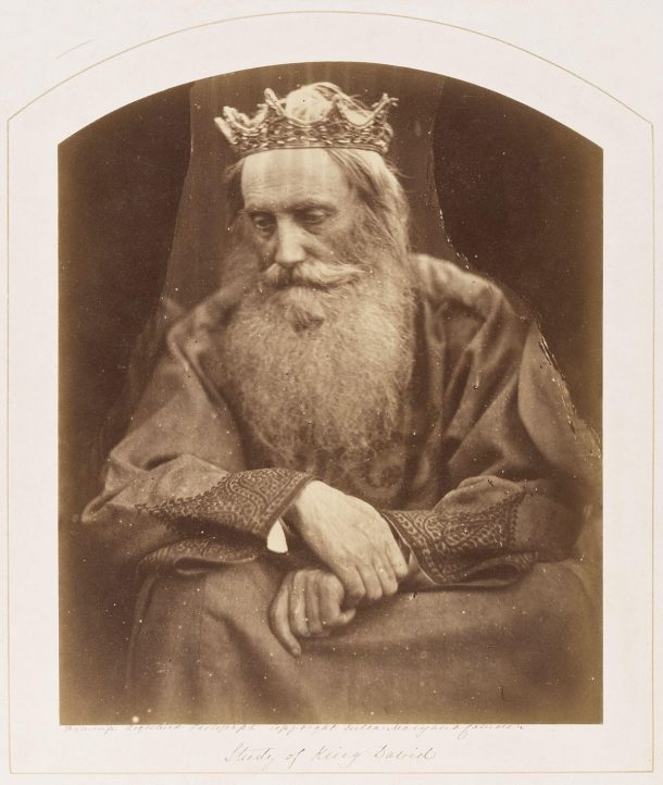 A bearded elderly man wearing a crown seated on a chair