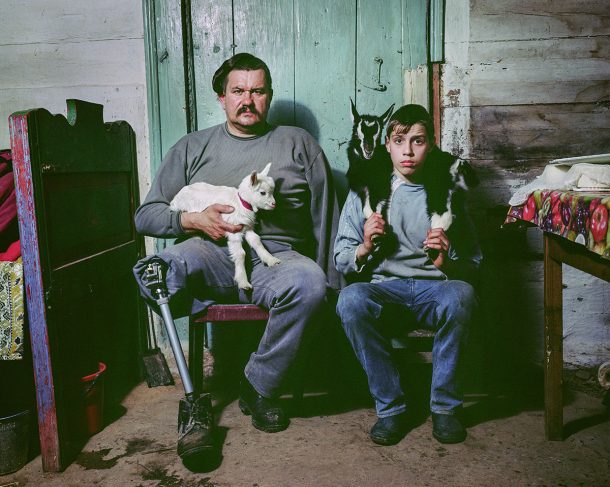 A man and a boy holding goats look at the camera. The man has a prosthetic right lower leg.