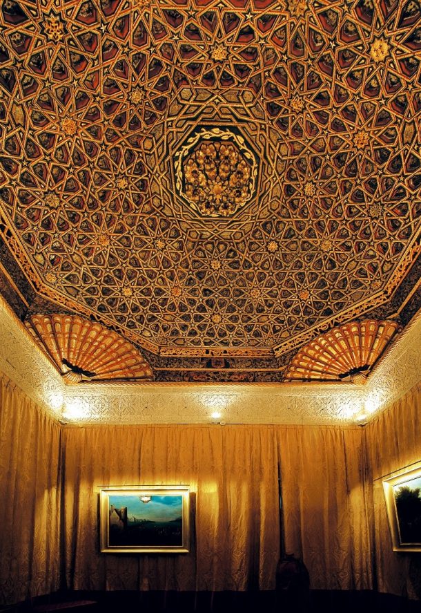 A highly decorated ceiling, supported by plaster cornicing and above walls covered by curtains