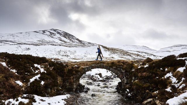 Jamie Kunta of Lonely Mountain Skis photographed by Alun Callend. Jamie walks across a stone bridge with skis over his shoulder in the Scottish Highlands.