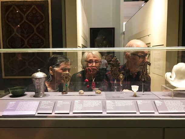Three people looking at a variety of objects through a glass cabinet in a gallery