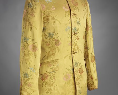 Nehru style jacket, characterised by the stand collar and long line cut. This jacket is made from a synthetic woven brocade fabric, with a yellow ground and floral motif in blue, pink and green. The jacket buttons from neck to waist with four self-covered buttons.