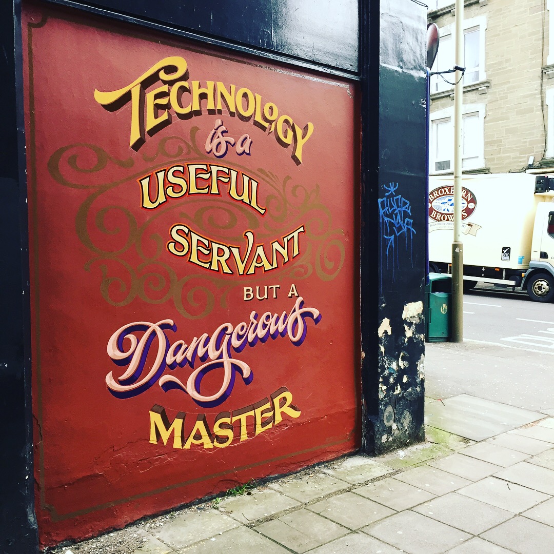 A painted sign in a street. The words read "Technology is a useful servant but a dangerous master" in beautiful, flowing typography.