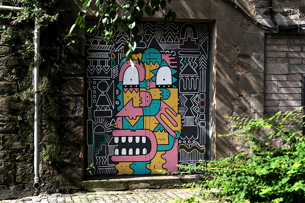 A painted door in an alleyway. The door iis yellow, pink and green and is a graphic and surreal looking face.