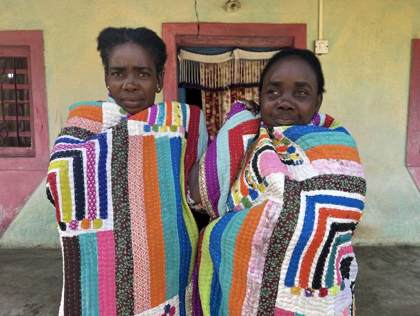 Two women wrapped in brightly coloured quilts outside a house
