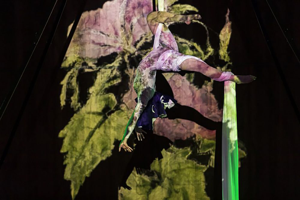 An acrobat wrapped in cloth, hanging suspended from a ceiling. Light projections cover her.
