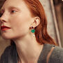 A red haired woman wears a heart shaped green earring with a freshwater pearl pendant.
