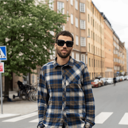 A white young man wearing a check shirt and black sunglasses stands on a city road.