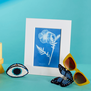 A cyanotype print featuring a white impression of a poppy flower with a white mount. The print is set on a blue table and background, together with a pair of yellow sunglasses and blue hairclips.