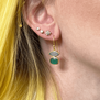 A blonde woman wearing gold hook earrings with green stones. 