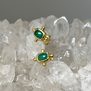A pair of stud gold earrings with a rectangular green stone in the centre, lying on a surface of white crystals.