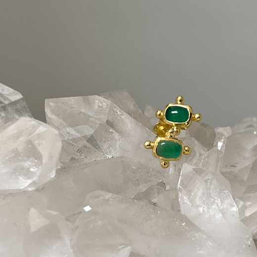 Two small stud gold earrings with a rectangular green stone in the centre, lying on a surface of white crystals.