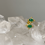 Two small stud gold earrings with a rectangular green stone in the centre, lying on a surface of white crystals.