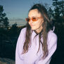 A young woman sitting outdoors wearing a pair of sunglasses and a lilac hoodie.