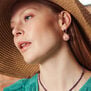 A white model with blonde hair wearing an earring featuring a round pink marbled stone pendant and a straw hat.