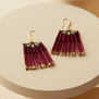 A pair of burgundy fan shaped earrings with gold hooks are laid on a cream circular prop..