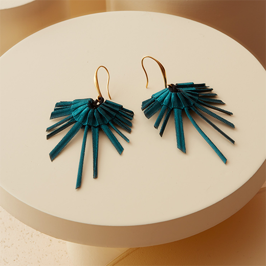 A pair of teal textile fan shaped earrings with gold hooks are laid on a beige circular prop.