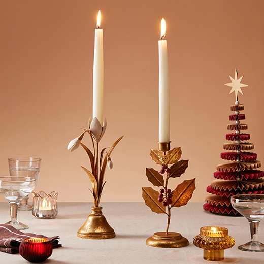 Two metal candlesticks in the form of a festive holly and branch and a snowdrop flower sit atop of a festive Christmas table.