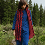 A young woman with brown hair modelling a long woollen scarf made with pink and burgundy yarns. She is wearing a blue jacket and is walking in an open field. 
