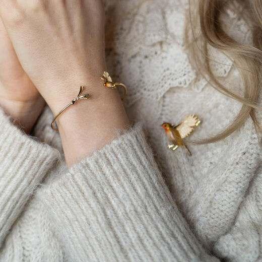 A woman wears a bracelet and a brooch featuring red-breasted flying robins. The photo shows a close up of her wrist and shoulder.
