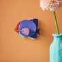 A wall art paper fish hung on an orange wall. A light blue vase with a pink flower can be seen in the foreground.
