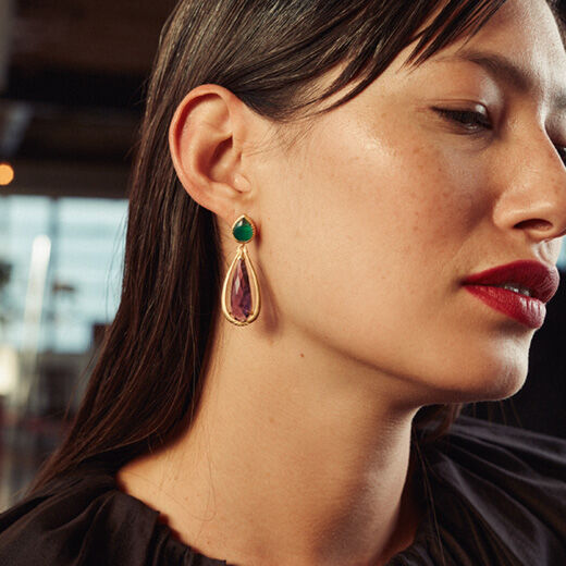 A young model with dark hair wears an earring featuring a gleaming green onyx at the base and teardrop shaped purple amethysts.