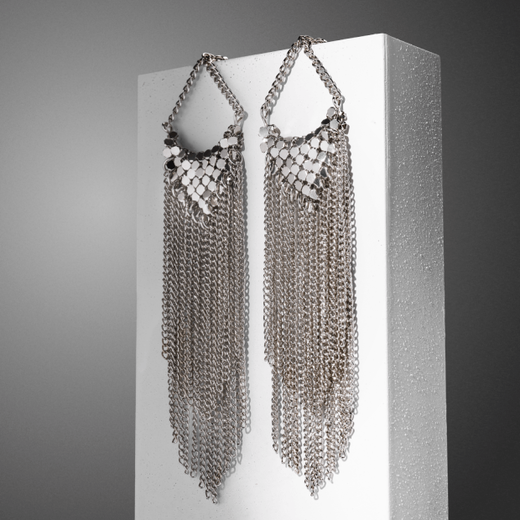 A pair of silver chainmail earrings hanging from a white block propped against a grey background.