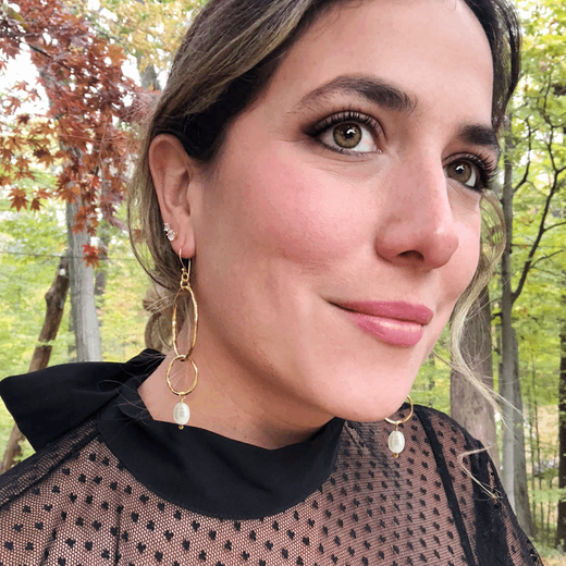 A woman with dark hair and black top wearing a pair of gold hoop earrings with a pendant pearl.
