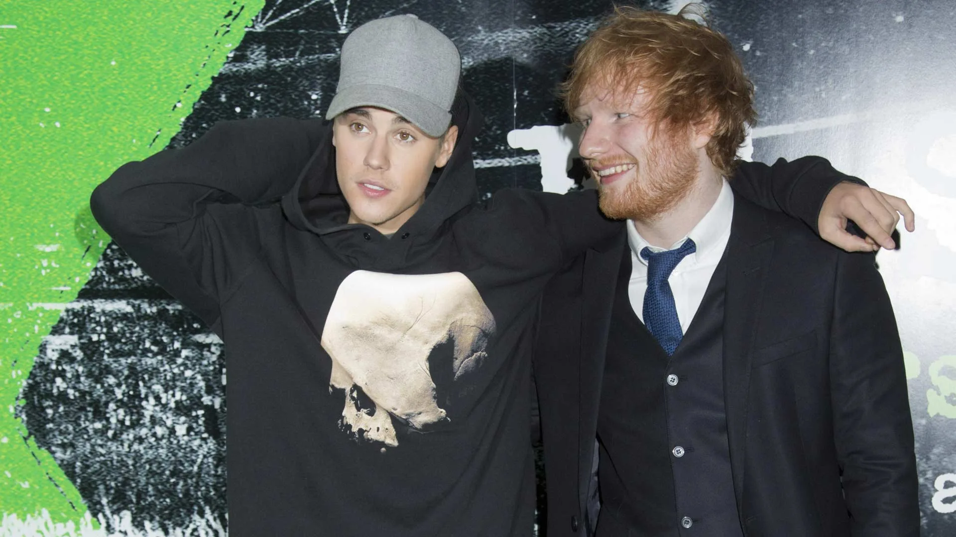 Justin Bieber and Ed Sheeran smiling and standing against a white, black and green wall.