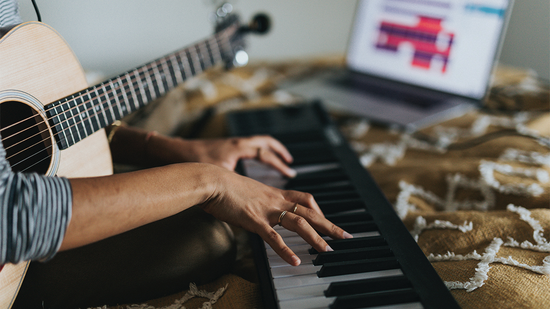 A person sat on a bed, playing a keyboard with a guitar on their lap