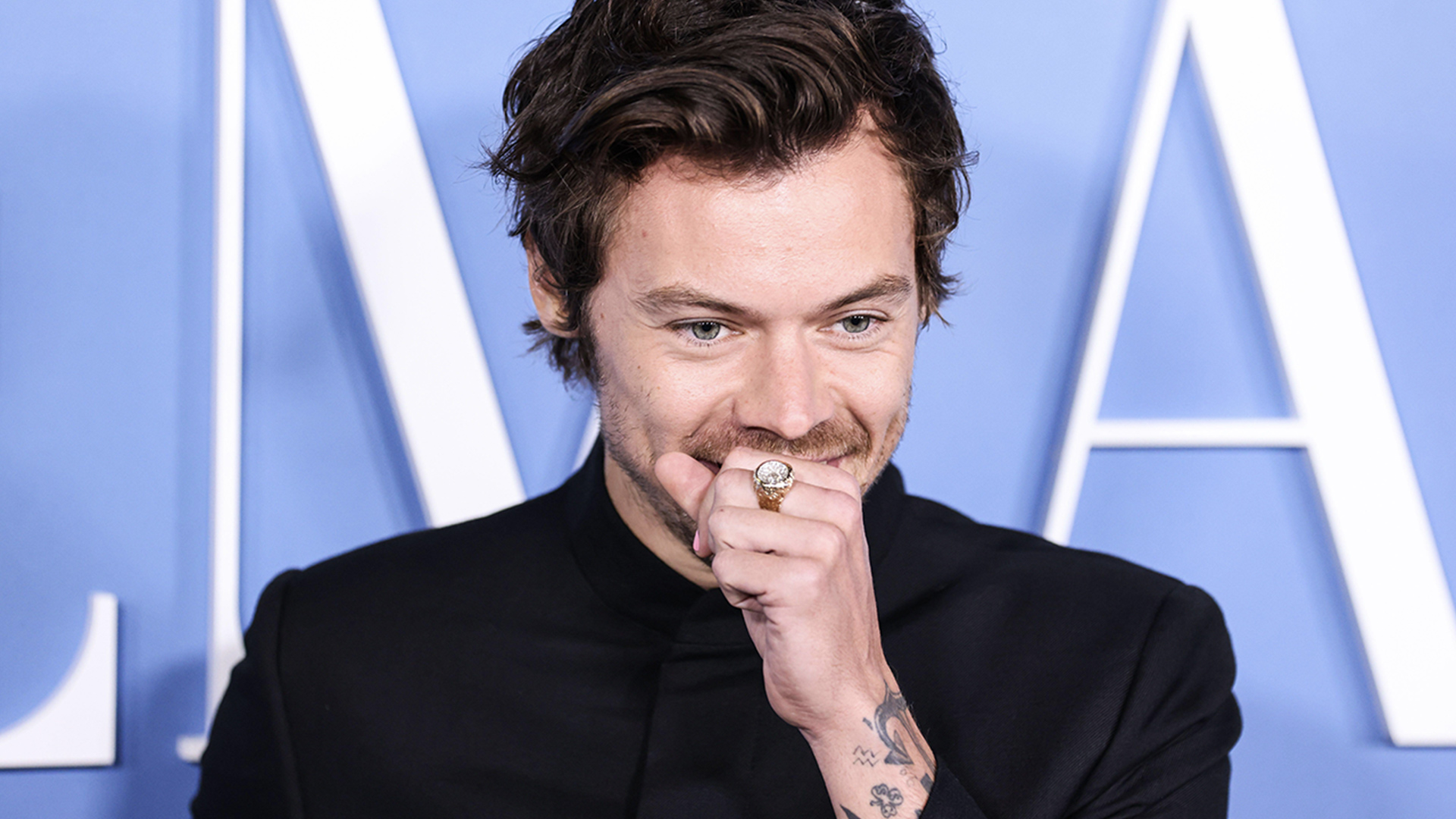Harry Styles with his hand to his mouth in a fist, as if he's covering a cough with a blue background