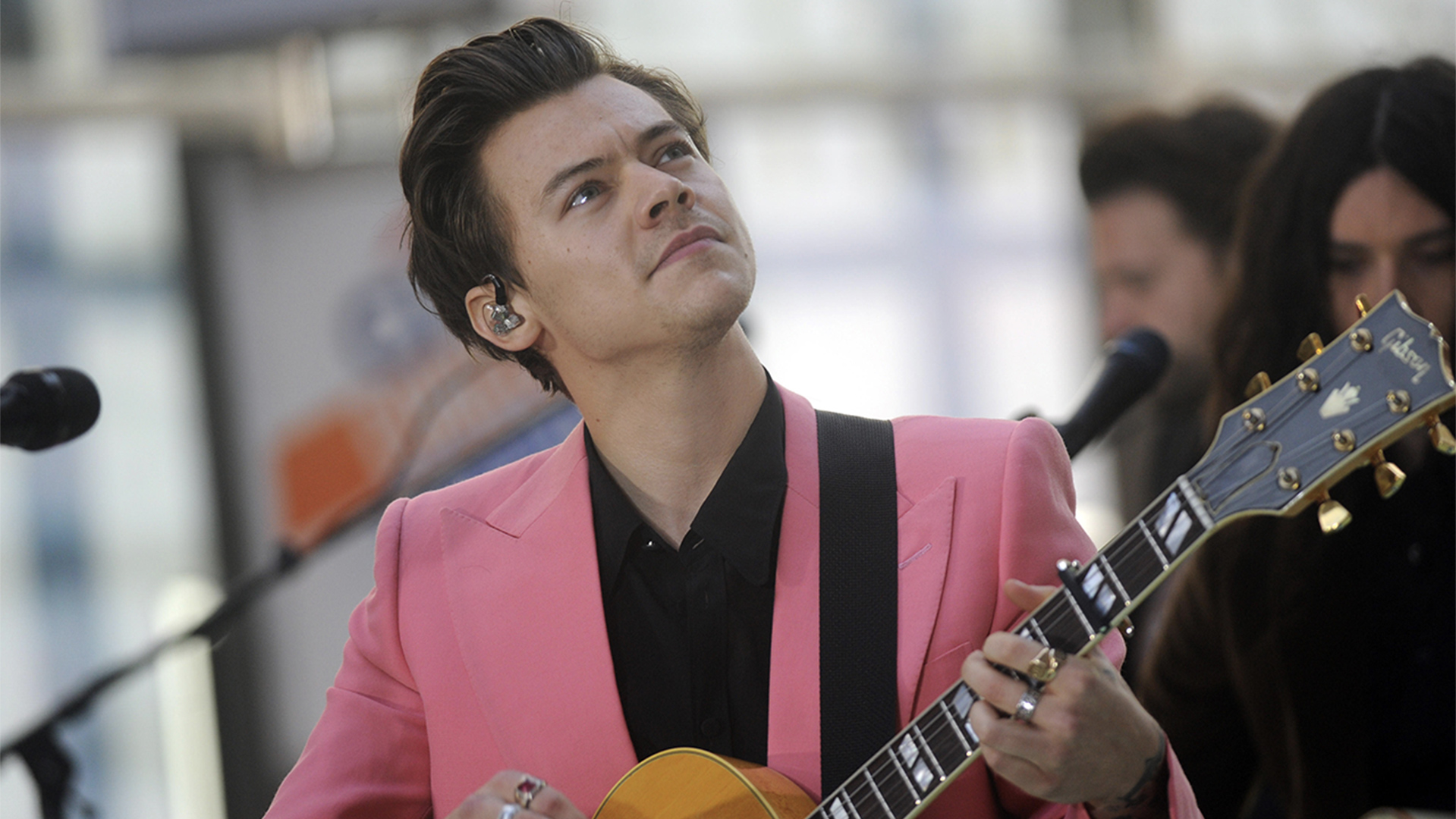 Harry Styles wearing a pink suit, holding a guitar and performing