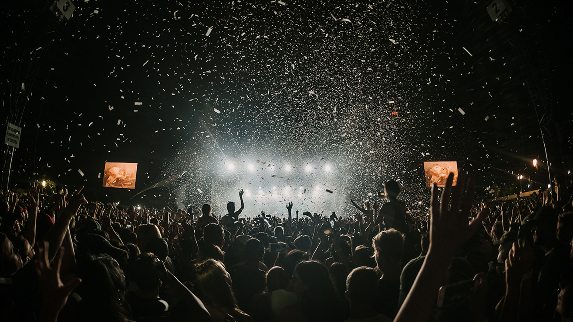 Large crowd with their hands up in the air at a festival at night, bright lights coming from the stage and confetti falling onto the crowd