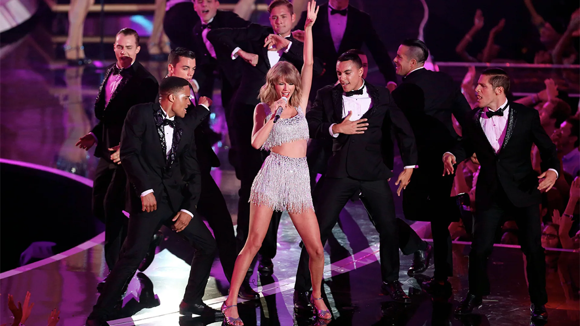 Tayor Swift performing on stage in silver glittery dress, holding silver glittery matched microphone, surrounded by male dancers dressed in black tuxedos.