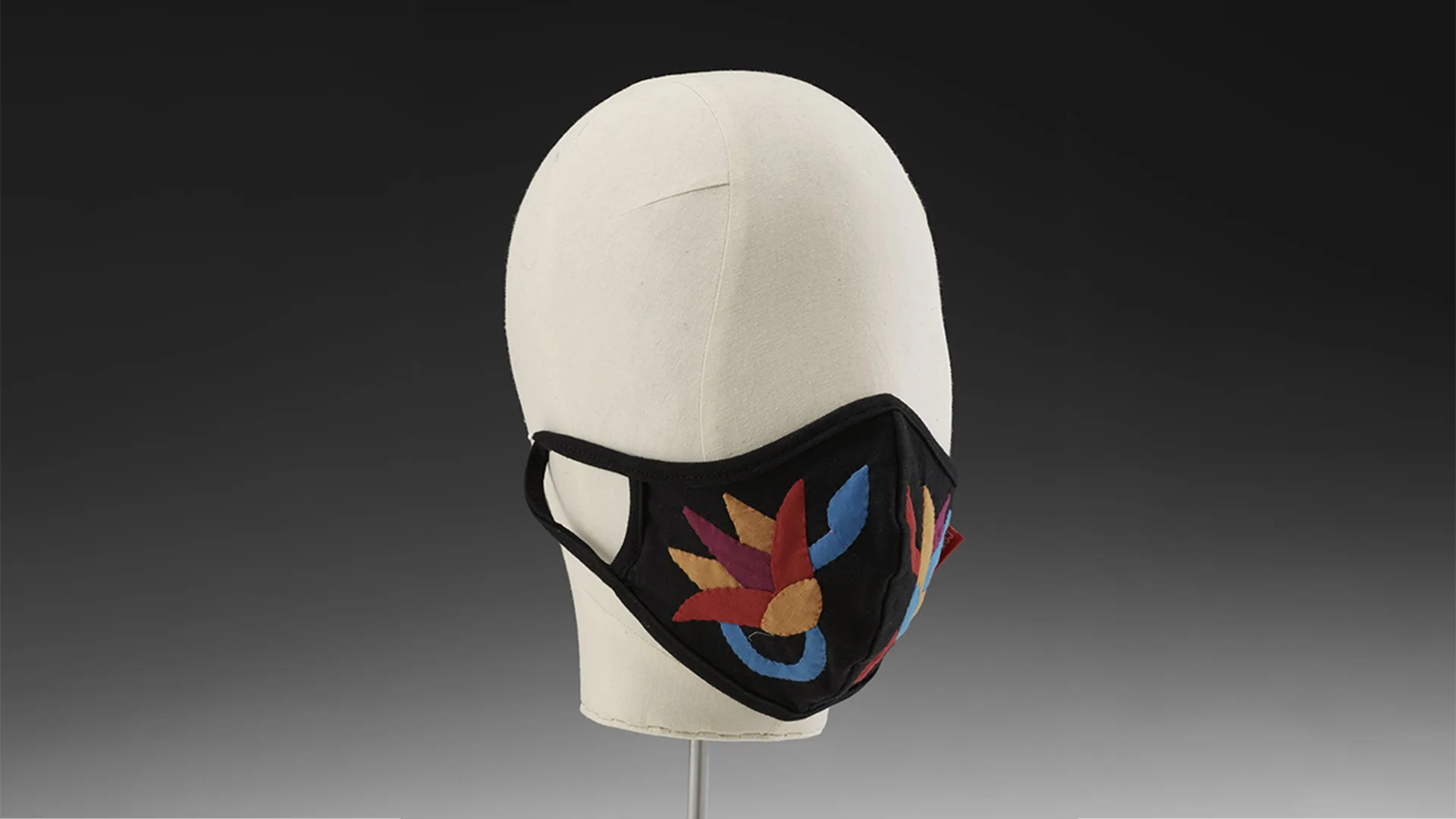 Mannequin head wearing black fabric mask decorated with floral pattern on black gradient background.