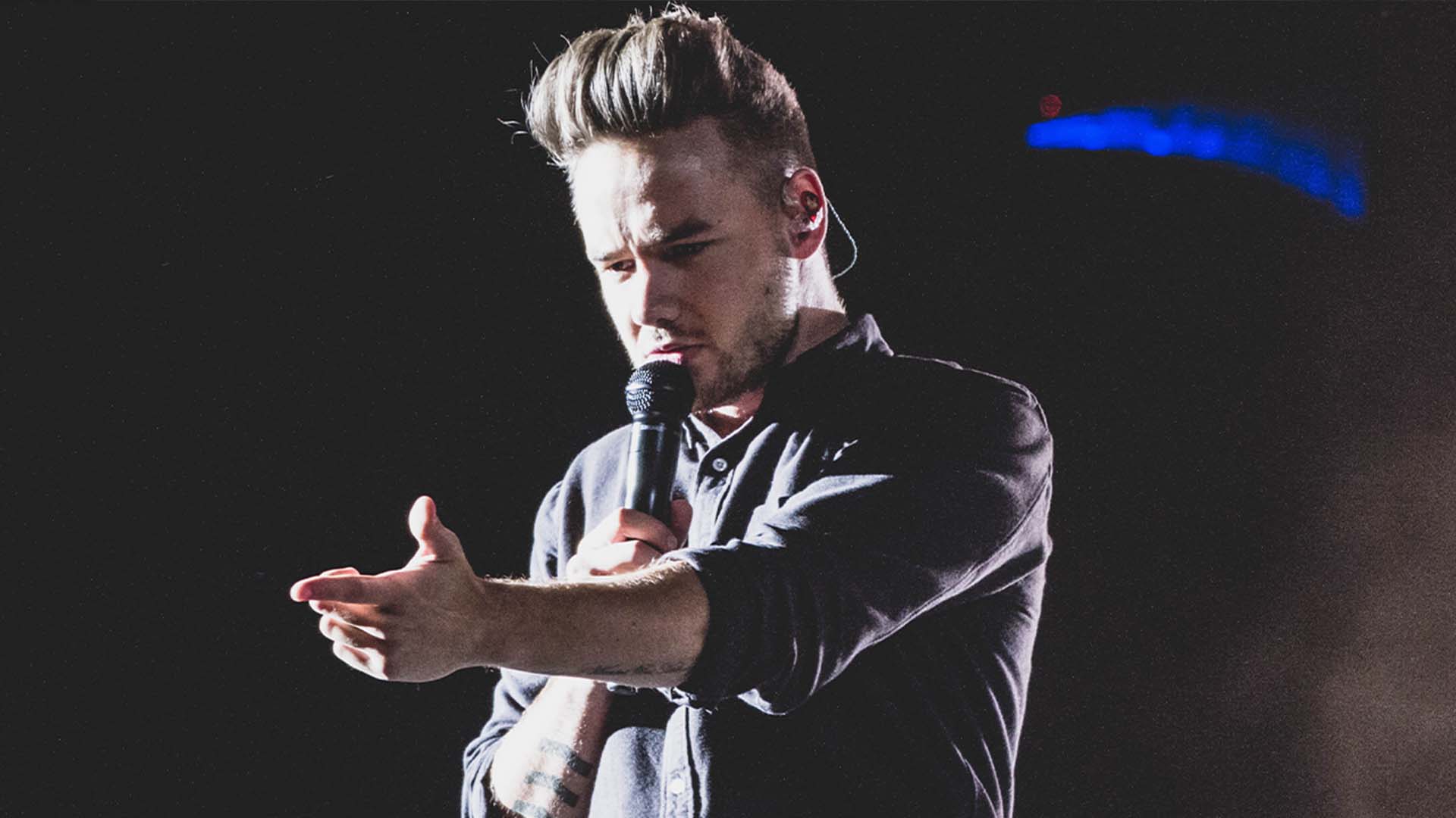 Liam Payne wearing black shirt, holding microphone while performing live on stage.