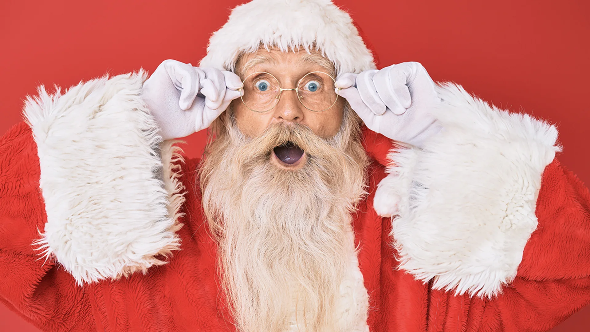 Close-up of Santa Claus in traditional red and white attire holding glasses with his mouth open.