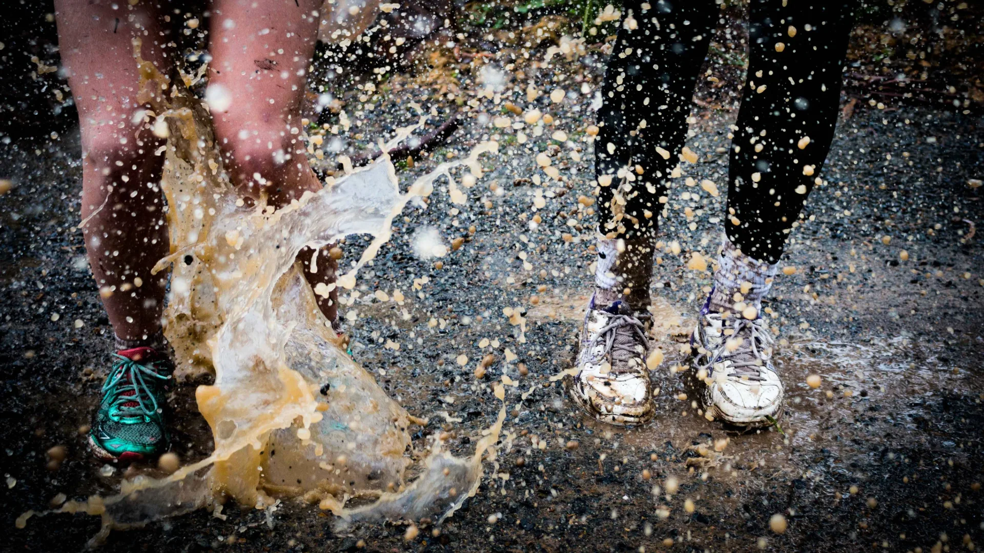 Two people wearing trainers jumping in a muddy puddle