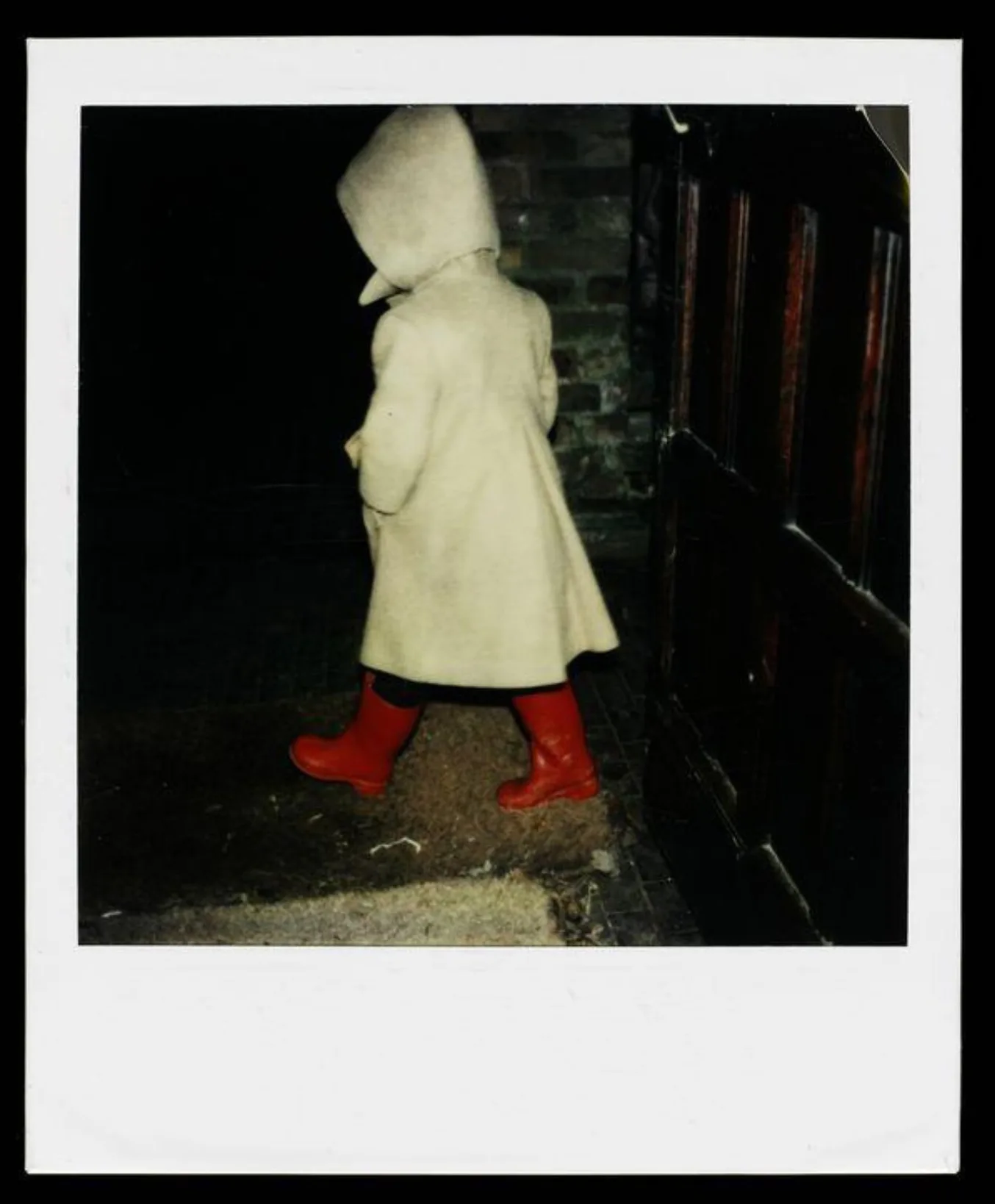 Polaroid depicting a child walking away from the camera wearing a white coat with hood up and red wellies
