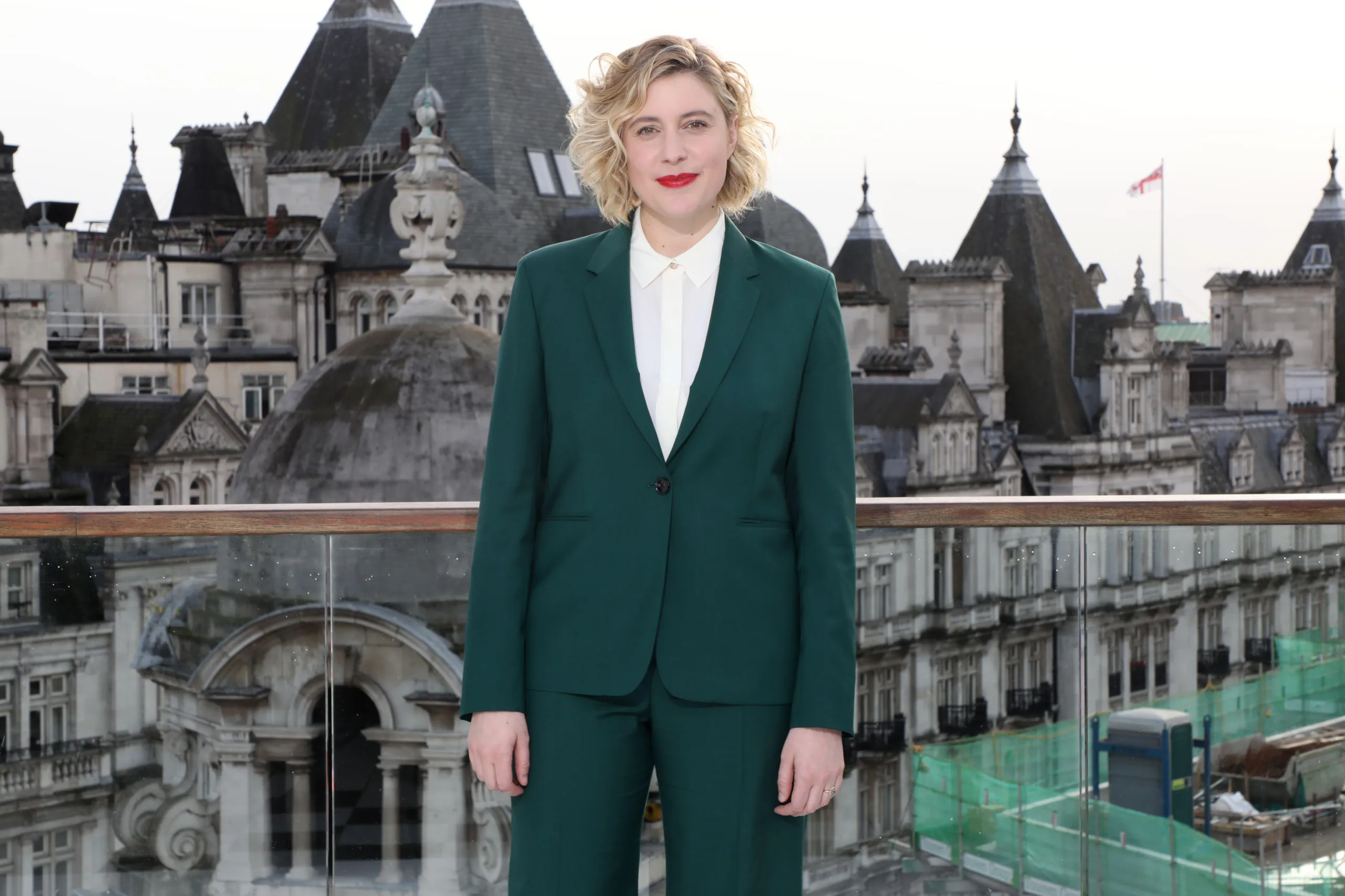 Greta Gerwig stands against city backdrop wearing green suit