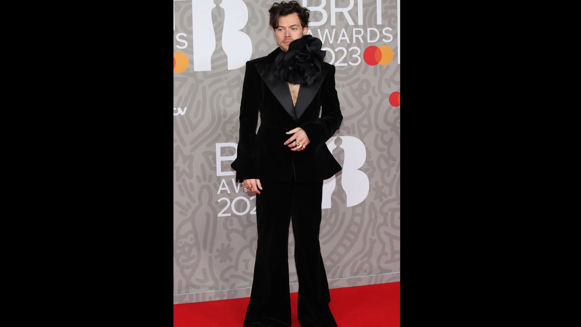 Photograph of Harry Styles wearing a black suit with an oversized flower choker on the red carpet at the BRIT awards