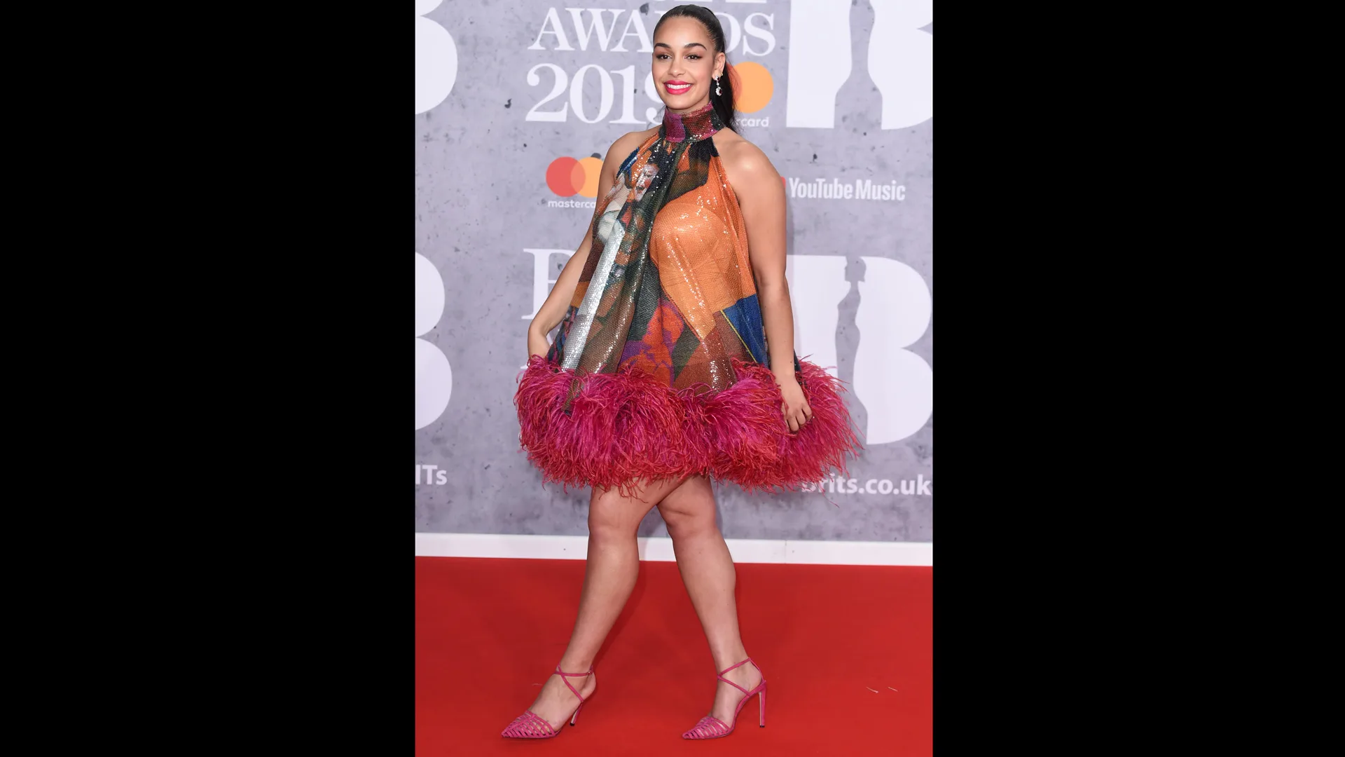 Photograph of Jorja Smith in a multi-coloured dress with red feather trim on the red carpet at the BRIT Awards