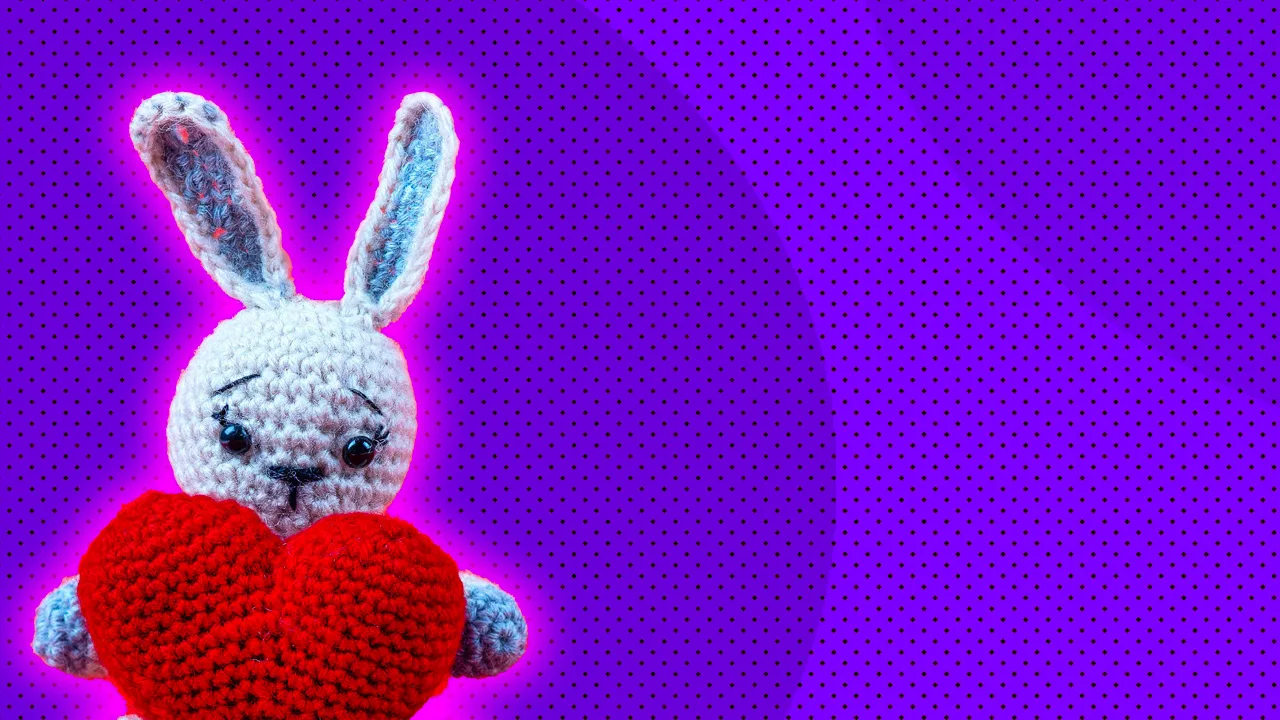 Photograph of a knitted bunny holding a heart against a purple dotted background with a pink halo