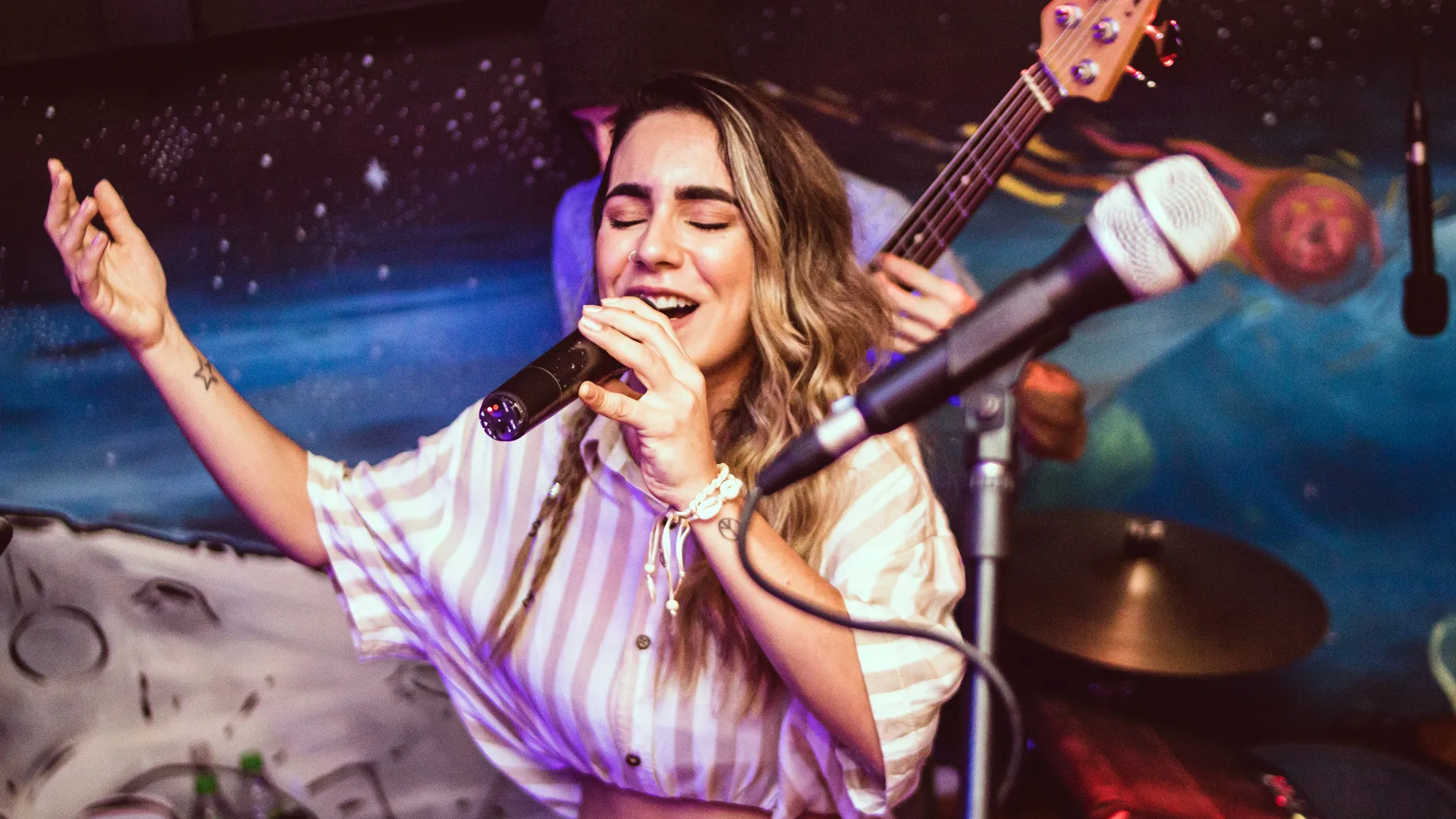 A photograph of a woman singing into a microphone with her eyes closed wearing a white striped top