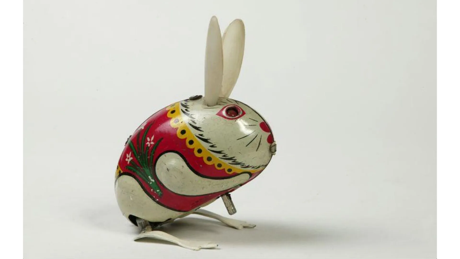 A photograph of a tin plate clockwork toy of a rabbit with a colourful jacket and painted red and black eyes, holding it's paw up to it's chin, against a white and grey background.