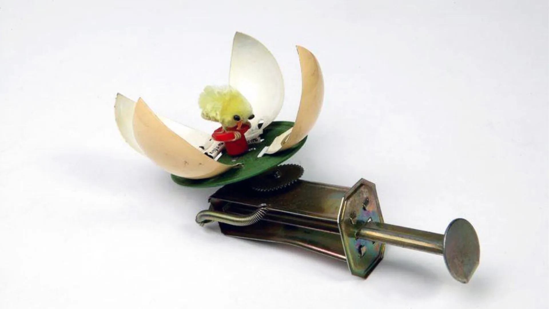 A photograph of a mechanical spinning toy with a plunger mechanism. The object consists of a segmented egg with a yellow chick inside, against a grey white gradient background.