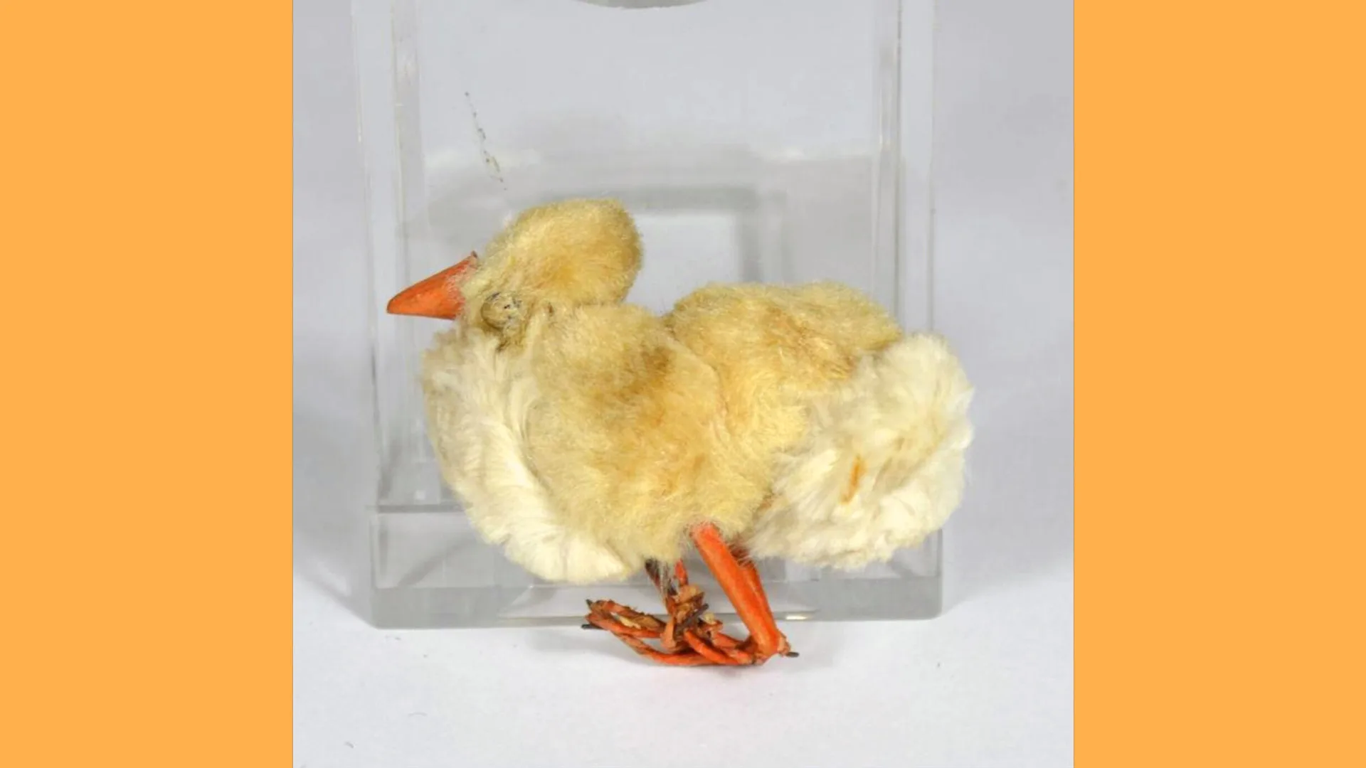 A photograph of a soft toy of a yellow chick with orange beak and feet. It is missing an eye. Against a grey background.
