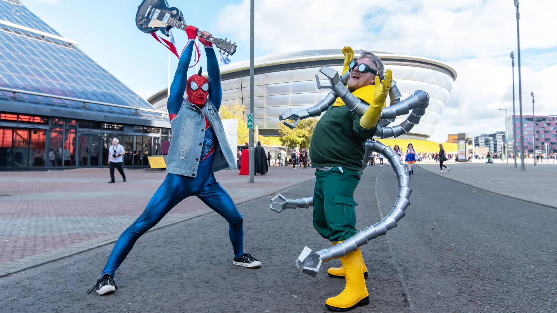 A photograph of two people at the Glasgow Comic Con outside in the carpark, dressed as Spiderman and Doctor Octopus pretending to have a fight as Spiderman holds a guitar up in the air and Doc Oc pretends to looks scared